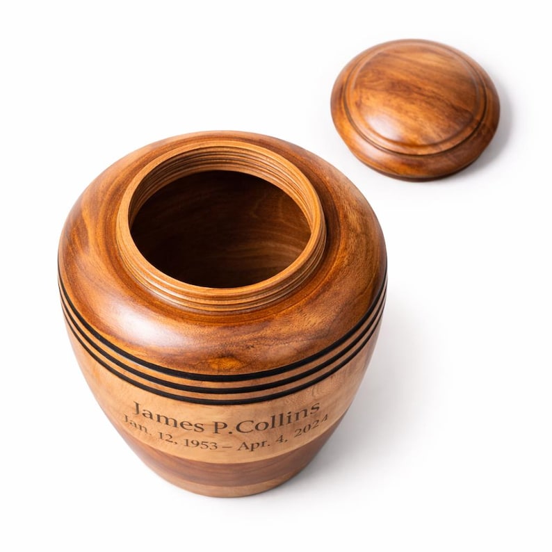 May include: A wooden urn with a removable lid. The urn is a warm brown color with black accent bands. The text "James P. Collins Jan. 12, 1953 - Apr. 4, 2024" is engraved on the side. The urn is designed to hold ashes.