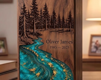 May include: A rectangular wooden urn featuring a laser-engraved forest scene with pine trees above a flowing river made of turquoise resin with gold leaf accents. The text "Oliver James" and "1985 – 2023" is engraved on the wood surface.