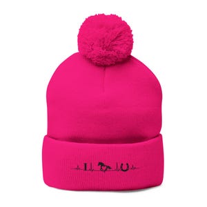 May include: A bright pink beanie hat with a fluffy pom-pom. The folded cuff displays a black graphic of a horse, horseshoe, and heartbeat line. This winter hat is a stylish accessory for horse riding fans.