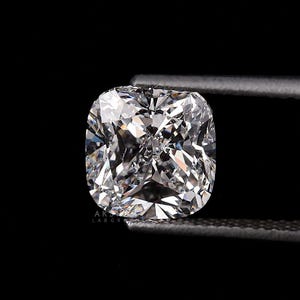 May include: A brilliant-cut, cushion-shaped diamond, held by a pair of tweezers against a black background. The diamond's facets reflect light, creating a sparkling effect. The diamond is a square shape with rounded corners.