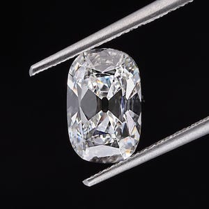 May include: A clear, cushion-cut diamond held by silver tweezers against a black background. The diamond's facets reflect light, creating a brilliant sparkle. The shape is rectangular with rounded corners, showcasing its clarity and cut.