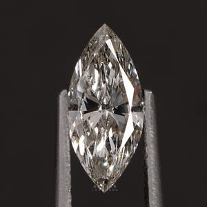 May include: A marquise-cut diamond held by tweezers against a black background. The diamond is clear with internal facets that reflect light. The shape is elongated with pointed ends, typical of this cut.