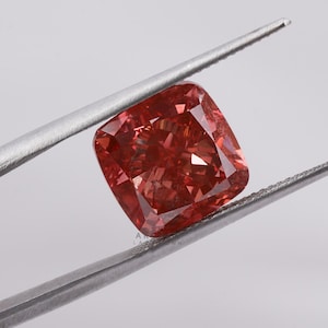 Op de afbeelding: Een kussenvormige, rood-oranje diamant wordt door een zilveren pincet tegen een witte achtergrond gehouden. De facetten van de diamant reflecteren licht, wat de schittering benadrukt. De vierkante vorm is licht afgerond.