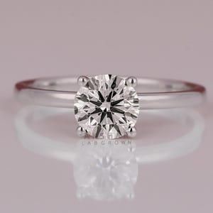 Puede incluir: Un anillo de compromiso solitario con un diamante de talla redonda. El diamante está engastado en un engaste de cuatro puntas sobre una fina banda de plata pulida. El anillo se muestra sobre una superficie reflectante.