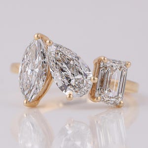 May include: A gold ring featuring three clear, sparkling diamonds: a marquise cut, a pear cut, and an emerald cut. The diamonds are set in a gold band, creating a luxurious and elegant design. The ring is displayed on a reflective surface.