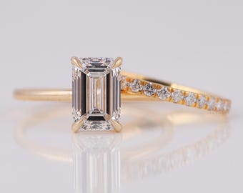 Op de afbeelding: Een gouden verlovingsring met een smaragdgeslepen diamant en een bijpassende band met kleine diamanten. De ringen staan tegen een witte achtergrond, die het licht reflecteert en een heldere, elegante uitstraling creëert.