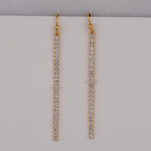 Può includere: Un paio di orecchini pendenti color oro. Ogni orecchino presenta una sottile barra verticale adornata da una fila di piccole pietre preziose trasparenti. Gli orecchini hanno un design semplice ed elegante, adatto a varie occasioni.