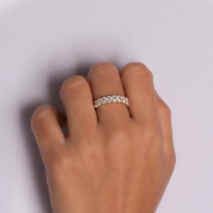 May include: A gold ring featuring multiple round, clear gemstones. The ring is worn on a finger against a pale purple backdrop. The gemstones sparkle, reflecting light.