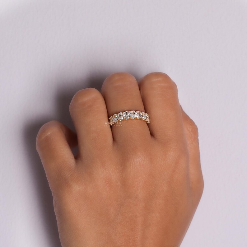 May include: A gold ring featuring multiple round, clear gemstones. The ring is worn on a finger against a pale purple backdrop. The gemstones sparkle, reflecting light.