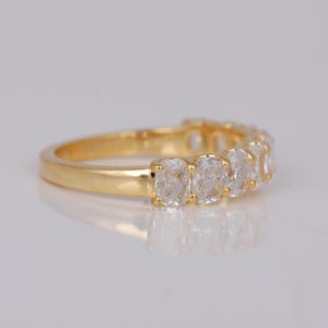 May include: A gold ring with a series of clear, cushion-cut gemstones. The ring's band is polished gold, and the gemstones are set closely together, creating a continuous sparkle. The ring is displayed on a light-coloured surface.