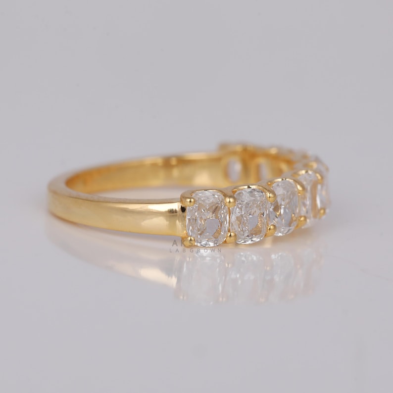 May include: A gold ring with a series of clear, cushion-cut gemstones. The ring's band is polished gold, and the gemstones are set closely together, creating a continuous sparkle. The ring is displayed on a light-coloured surface.