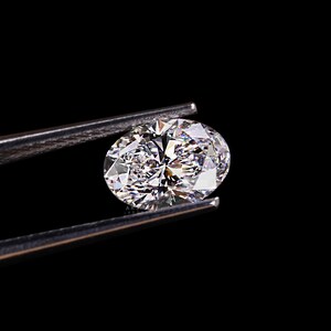 May include: A sparkling, oval-cut diamond held by silver tweezers against a black background. The diamond's facets reflect light, creating a brilliant display of color and sparkle. The image highlights the clarity and cut of the gemstone.