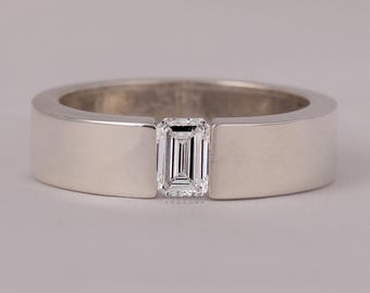 May include: A silver ring with a rectangular, emerald-cut diamond set in the center. The band is wide and flat, with a polished finish. The diamond is clear and reflects light.