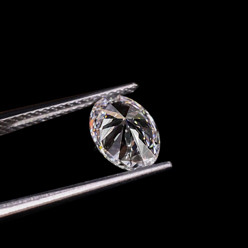 May include: A brilliant, oval-cut diamond held by silver tweezers against a black background. The diamond's facets reflect light, creating a sparkling effect. The image highlights the clarity and brilliance of the gemstone.