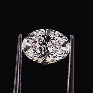 May include: A brilliant, oval-cut diamond held by a pair of silver tweezers against a black background. The diamond sparkles with numerous facets, reflecting light and creating a dazzling display. The image is a close-up, highlighting the gem's clarity and brilliance.