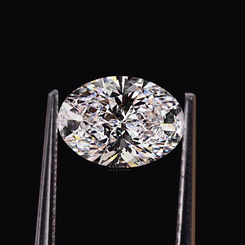 May include: A brilliant, oval-cut diamond held by a pair of silver tweezers against a black background. The diamond sparkles with numerous facets, reflecting light and creating a dazzling display. The image is a close-up, highlighting the gem's clarity and brilliance.