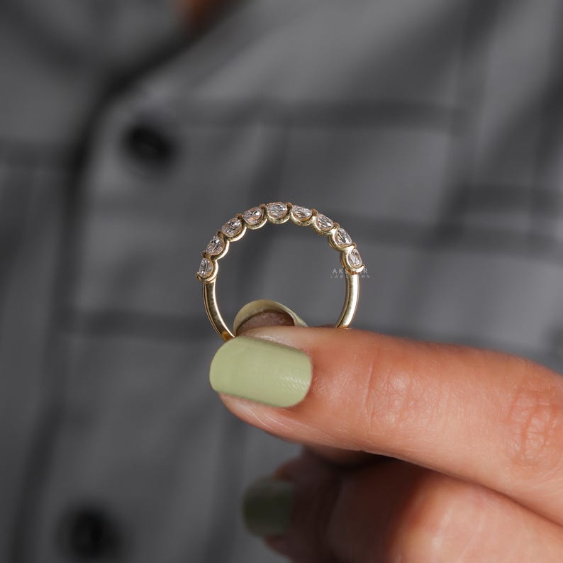 May include: A gold ring featuring a line of clear gemstones. The ring is held by a person's fingers with light green nail polish. The ring is a half-eternity style, with the gemstones set closely together.