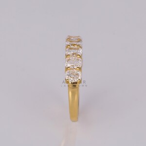 May include: A gold ring with a band set with a row of clear, square-cut gemstones. The ring's band is a bright gold colour, and the gemstones are set closely together. The ring is shown against a neutral background.