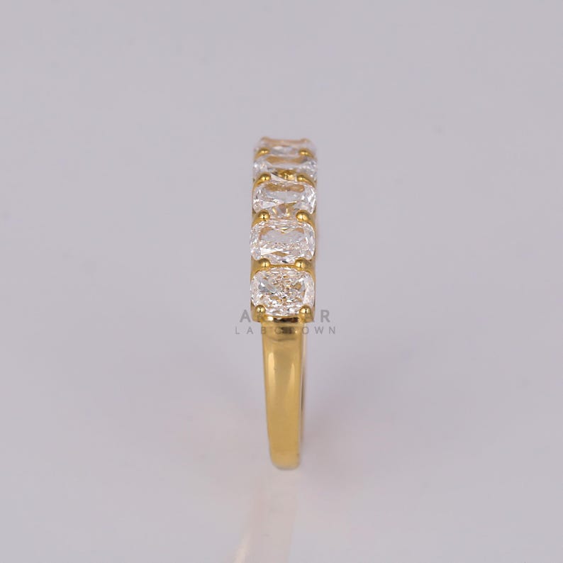 May include: A gold ring with a band set with a row of clear, square-cut gemstones. The ring's band is a bright gold colour, and the gemstones are set closely together. The ring is shown against a neutral background.
