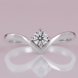 Könnte beinhalten: Ein silberner Verlobungsring mit einem runden, klaren Diamanten als Mittelstein. Der Ring hat ein V-förmiges Band, das sich nach oben wölbt, um den Diamanten zu treffen. Der Ring ist vor einem weichen rosa Hintergrund platziert.