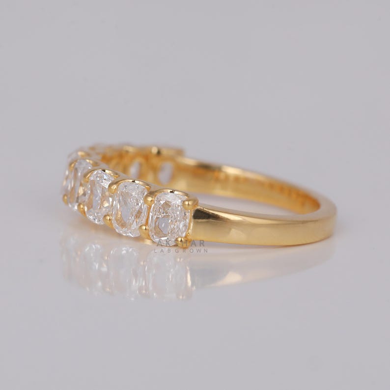 May include: A gold ring with a row of clear, oval gemstones. The ring's band is a polished gold colour, and the gemstones are set close together, creating a continuous line of sparkle. The ring is displayed on a reflective surface.