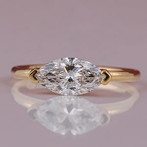 May include: A marquise-cut diamond ring with a gold band. The diamond is set horizontally, and the band has heart-shaped accents on either side of the diamond. The ring is displayed on a reflective surface.