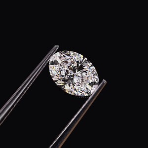 May include: A brilliant, oval-cut diamond held by a pair of tweezers against a black background. The diamond sparkles with numerous facets, reflecting light and creating a dazzling display of brilliance. The tweezers are silver-colored.