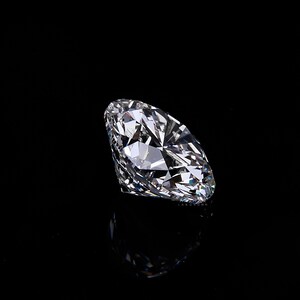 May include: A large, round-cut diamond with numerous facets, reflecting light and appearing brilliant. The gemstone is set against a black background, creating a dramatic contrast and highlighting its clarity and sparkle. The diamond's reflection is visible below.