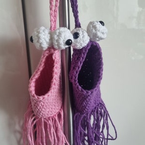 Könnte beinhalten: Zwei gehäkelte Hängetaschen mit Fransen, eine rosa und eine lila, jede mit zwei weißen gehäkelten Augen und schwarzen Pupillen.