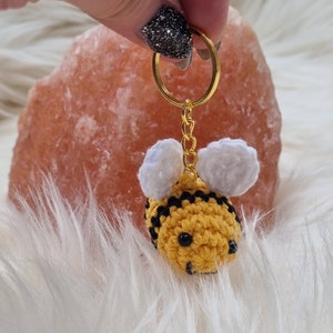 Könnte beinhalten: Ein gehäkelter Bienenschlüsselanhänger mit schwarzen und gelben Streifen, weißen Flügeln und schwarzen Augen. Die Biene ist an einem goldenen Schlüsselring befestigt.
