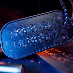 Może przedstawiać: Zbliżenie niebieskiej metalowej blaszki z wytłoczonymi słowami "STREAM OVERLAY". Blaszka jest przymocowana do łańcuszka kulkowego. Tło jest ciemne z pomarańczowymi i czerwonymi akcentami.
