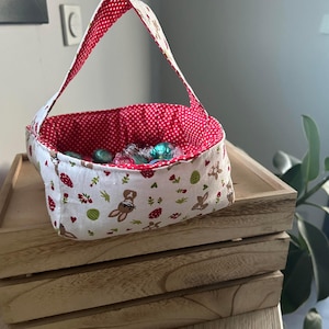 Puede incluir: Una cesta de Pascua de tela con interior de lunares rojos y exterior blanco estampado con conejitos, huevos y flores. La cesta contiene caramelos envueltos y está sobre una bandeja de madera.