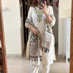 Può includere: Completo kurta ricamato bianco sporco con pantaloni abbinati e uno scialle stampato. Il kurta presenta ricami verdi intorno allo scollo e alle maniche. Lo scialle ha una stampa floreale e geometrica con frange. I pantaloni hanno finiture ricamate verdi.