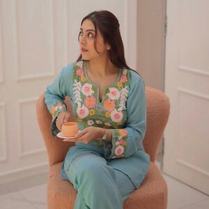 Op de afbeelding: Lichtblauw geborduurd tweedelig outfit met bloemmotieven in roze, wit en geel. De outfit bestaat uit een top met lange mouwen en een bijpassende broek. De persoon zit op een roze stoel en houdt een kleine oranje kop vast.