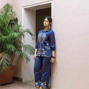 May include: A two-piece navy blue outfit featuring an embroidered top and matching pants. The top has a detailed floral design around the collar and down the front. The pants have floral embroidery at the hem. The outfit is worn with gold-colored earrings and sandals.