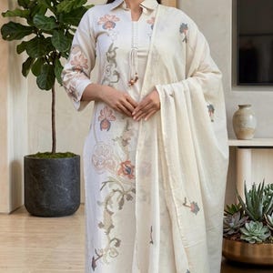 May include: An off-white long-sleeved tunic with a collared neckline and a front placket featuring hanging decorative beads. The tunic is adorned with intricate floral embroidery in muted orange, blue, and green tones, paired with matching wide-leg trousers.
