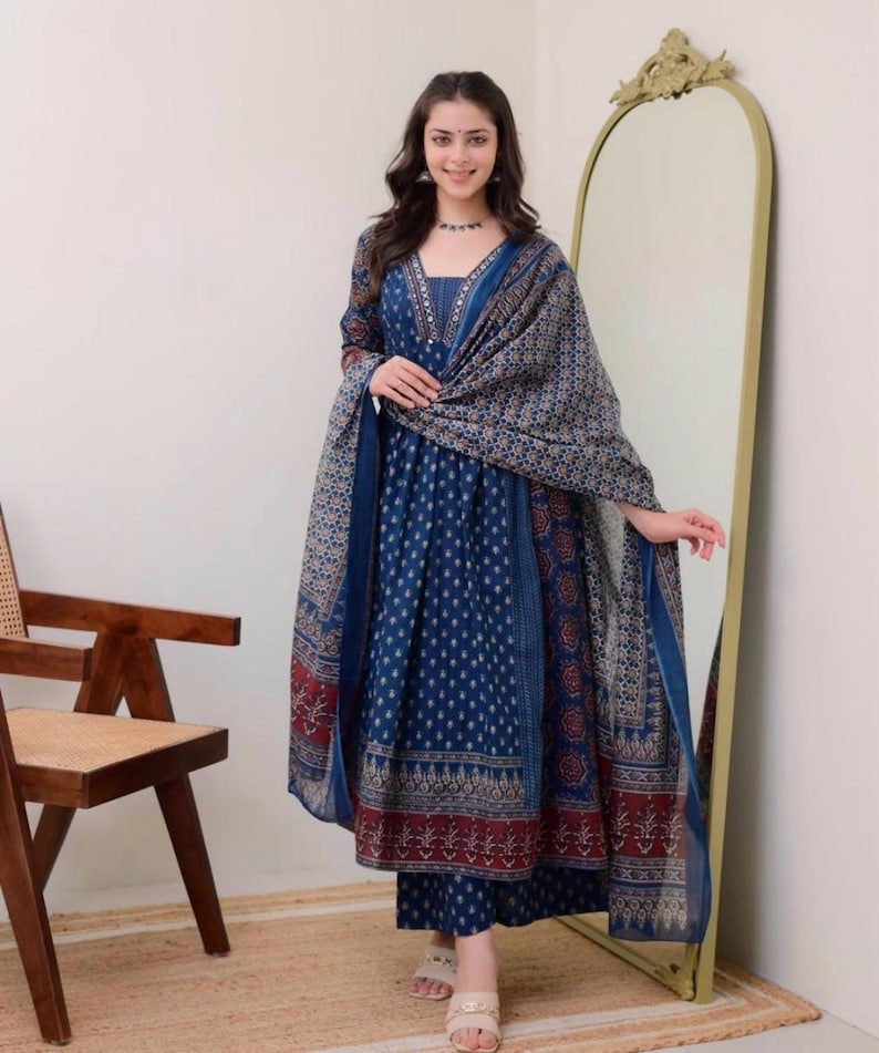 K&ouml;nnte beinhalten: Ein blaues bedrucktes Salwar Kameez-Set mit passendem Dupatta. Das Outfit hat einen V-Ausschnitt und lange &Auml;rmel. Der Stoff hat ein Muster aus kleinen wei&szlig;en Punkten und floralen Bord&uuml;ren. Die Person tr&auml;gt Sandalen.
