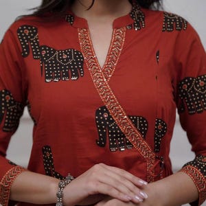 May include: Rust-colored wrap top with a black elephant print. The top features a V-neckline with gold sequin trim and long sleeves. The elephant design is repeated across the garment, creating a unique pattern.