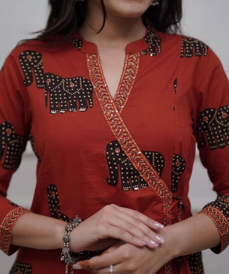 May include: Rust-colored wrap top with a black elephant print. The top features a V-neckline with gold sequin trim and long sleeves. The elephant design is repeated across the garment, creating a unique pattern.