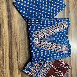 K&ouml;nnte beinhalten: Gefalteter blauer Stoff mit wei&szlig;en Blumenmustern und einem dekorativen Rand mit roten und wei&szlig;en Akzenten. Der Stoff ist auf einer Holzoberfl&auml;che angeordnet und zeigt das komplizierte Design und den Farbkontrast.