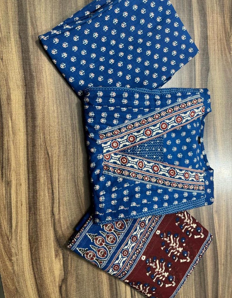 K&ouml;nnte beinhalten: Gefalteter blauer Stoff mit wei&szlig;en Blumenmustern und einem dekorativen Rand mit roten und wei&szlig;en Akzenten. Der Stoff ist auf einer Holzoberfl&auml;che angeordnet und zeigt das komplizierte Design und den Farbkontrast.