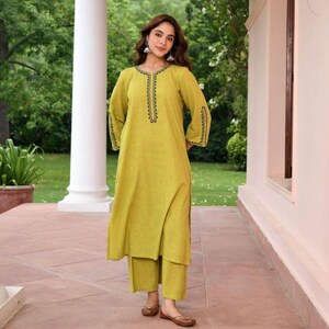 May include: A light green tunic and pants set. The tunic has long sleeves and a split hem, with black embroidered detailing around the neckline and cuffs. The pants are a matching solid color. The outfit is worn with brown flats.
