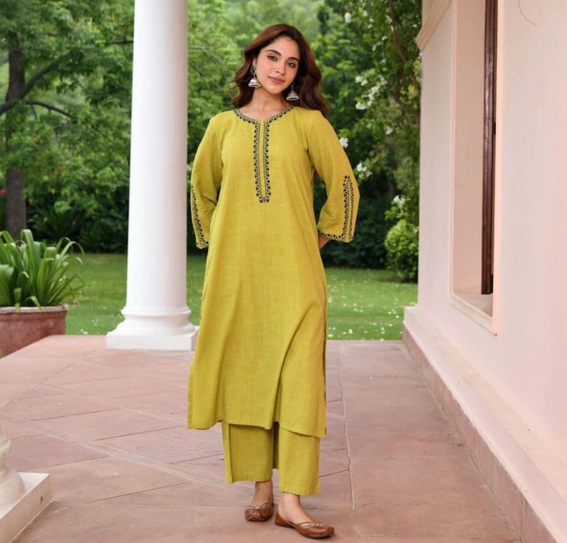 May include: A light green tunic and pants set. The tunic has long sleeves and a split hem, with black embroidered detailing around the neckline and cuffs. The pants are a matching solid color. The outfit is worn with brown flats.