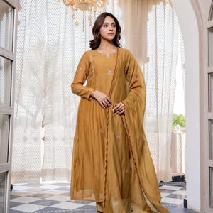 May include: Mustard-coloured, long-sleeved dress with delicate embroidery and matching scarf. The dress has a flowing silhouette and is paired with clear sandals. The background includes a checkered floor and ornate doorway.