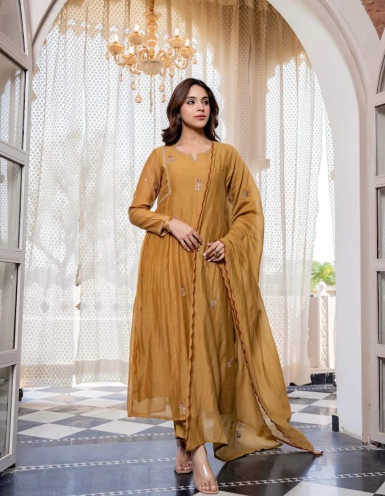 May include: Mustard-coloured, long-sleeved dress with delicate embroidery and matching scarf. The dress has a flowing silhouette and is paired with clear sandals. The background includes a checkered floor and ornate doorway.
