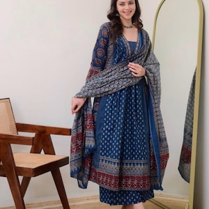 K&ouml;nnte beinhalten: Eine Frau in einem blau bedruckten indischen Anzug mit passendem Schal. Der Anzug hat ein detailliertes Muster in Blau-, Rot- und Beiget&ouml;nen. Das Outfit umfasst ein lang&auml;rmliges Oberteil, eine Hose und einen drapierten Schal.