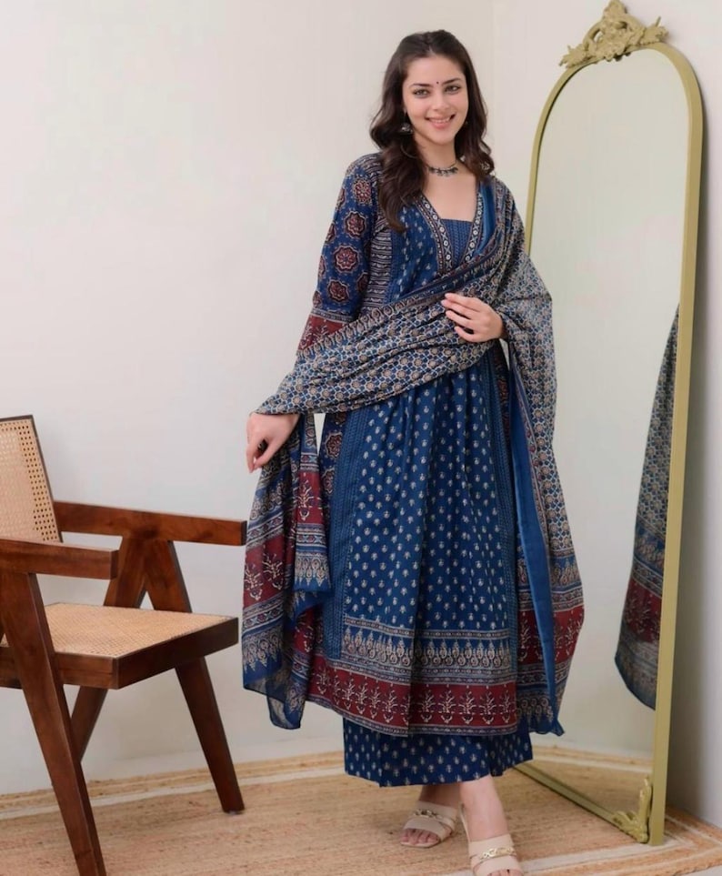 K&ouml;nnte beinhalten: Eine Frau in einem blau bedruckten indischen Anzug mit passendem Schal. Der Anzug hat ein detailliertes Muster in Blau-, Rot- und Beiget&ouml;nen. Das Outfit umfasst ein lang&auml;rmliges Oberteil, eine Hose und einen drapierten Schal.