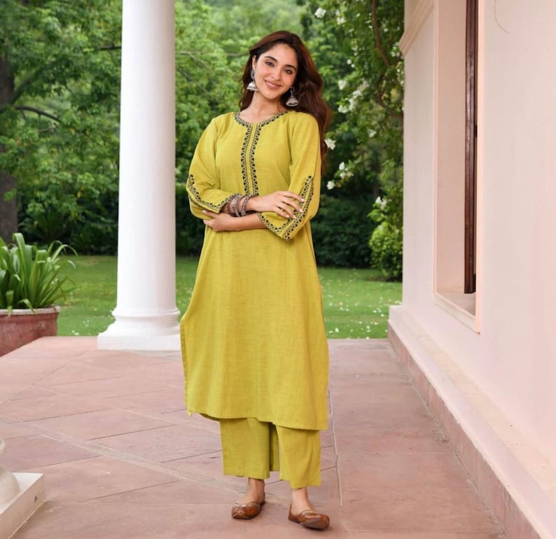 May include: A lime green tunic and matching pants set. The tunic features black embroidered detailing around the neckline and sleeves. The outfit is made of a lightweight fabric and is suitable for warm weather. The model is wearing brown flats and silver earrings.