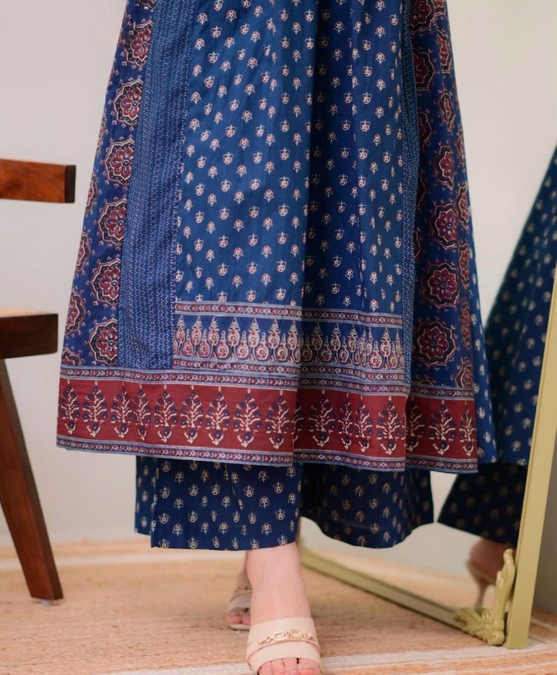K&ouml;nnte beinhalten: Ein blau gemusterter Rock und eine passende Hose mit dekorativem Rand. Der Rock und die Hose haben ein sich wiederholendes Blumenmuster in Wei&szlig; und Rot. Das Outfit wird mit beigen Sandalen kombiniert.