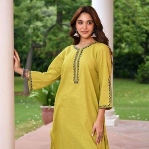 May include: A long-sleeved, chartreuse-colored tunic with black embroidered detailing along the neckline and sleeve cuffs. The tunic is made of a lightweight fabric and has a relaxed fit. The model is wearing silver earrings and bracelets.
